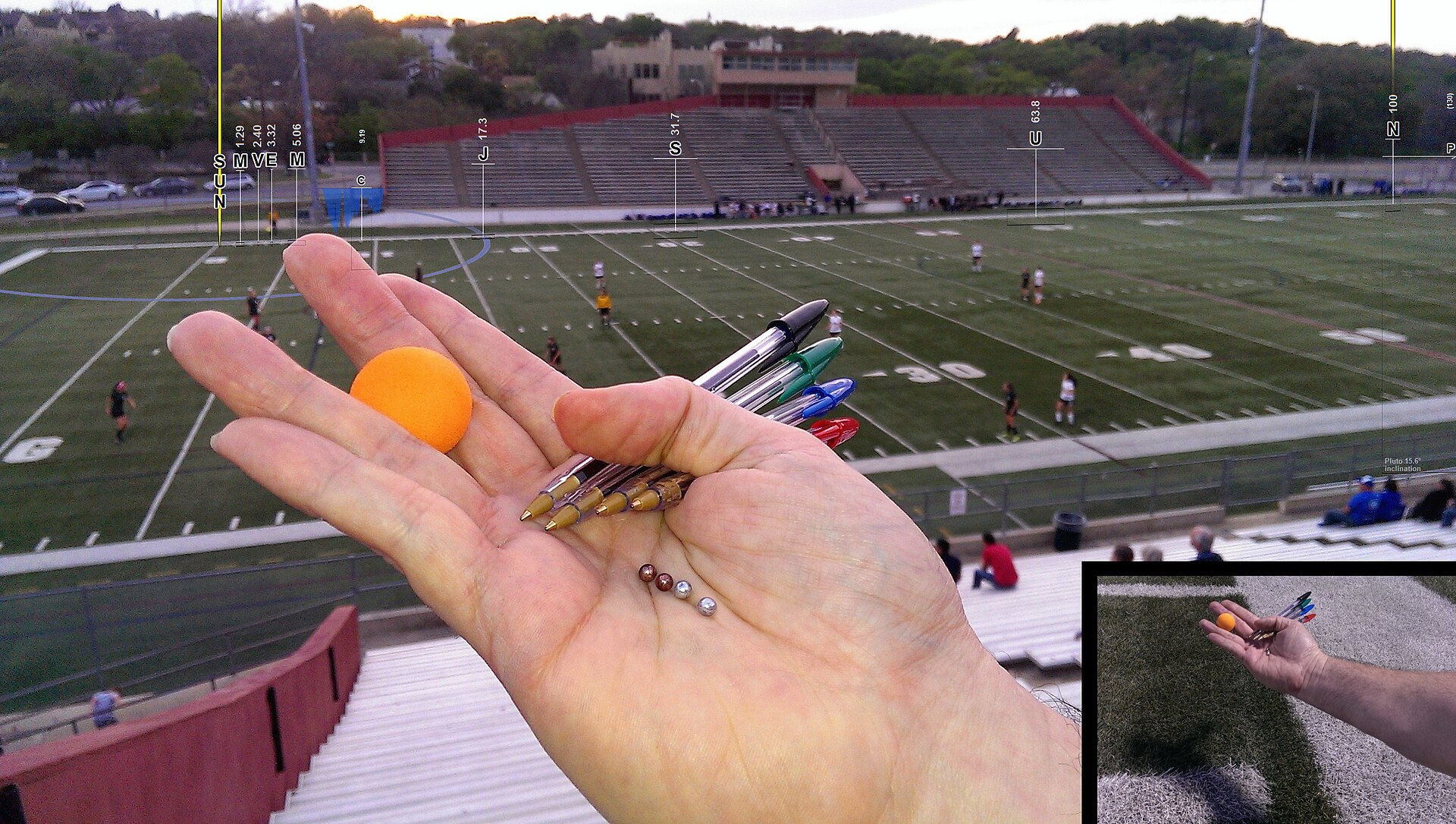 A hand holding a small ball, ballpoint pens, and BB pellets, in front of a football field