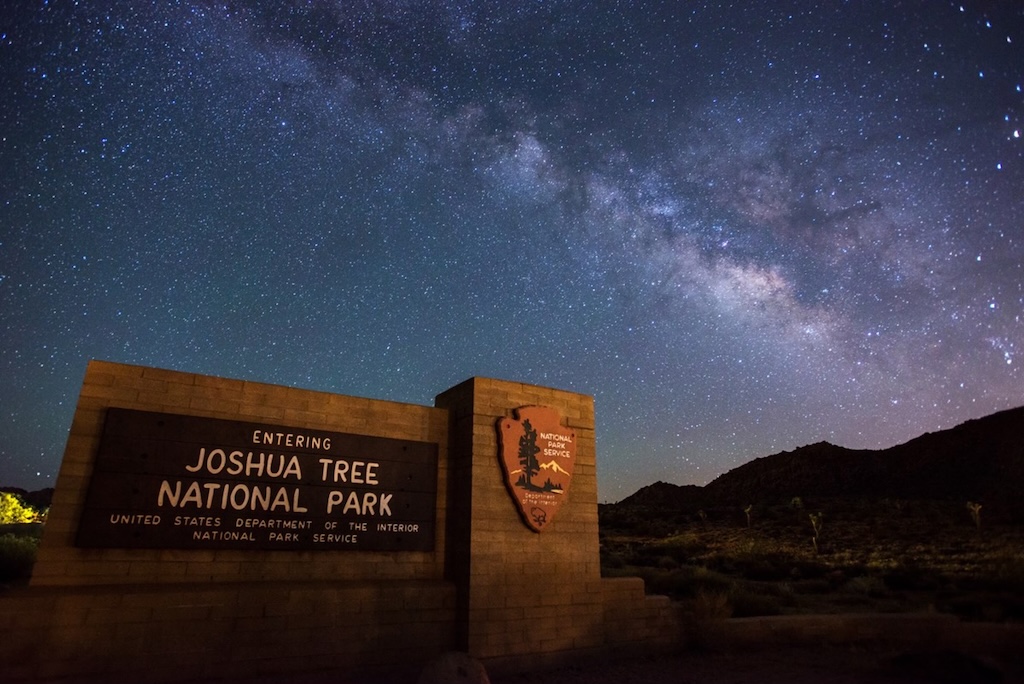 A night sky photo showing the Milky Way over a sign saying Entering Joshua Tree National Park