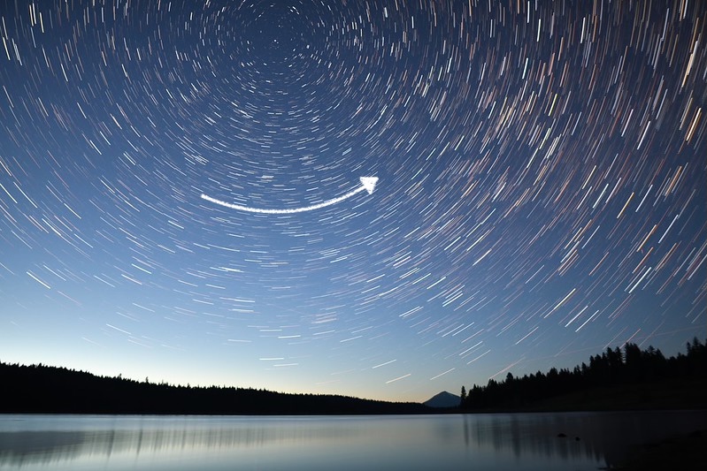 Star trails make circles high in the sky above a lake. An arrow below the center of the circles points to the right.