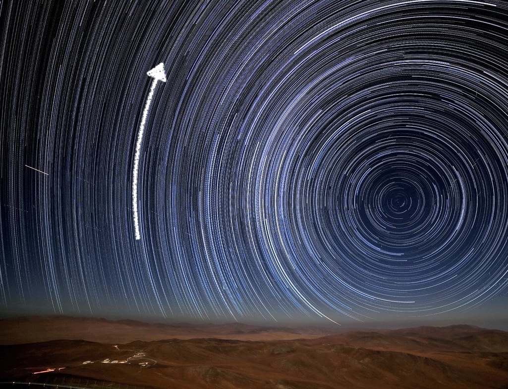 Star trails make circles around a point to the right of the image above a desert. An arrow to the left of the circles points up from the horizon